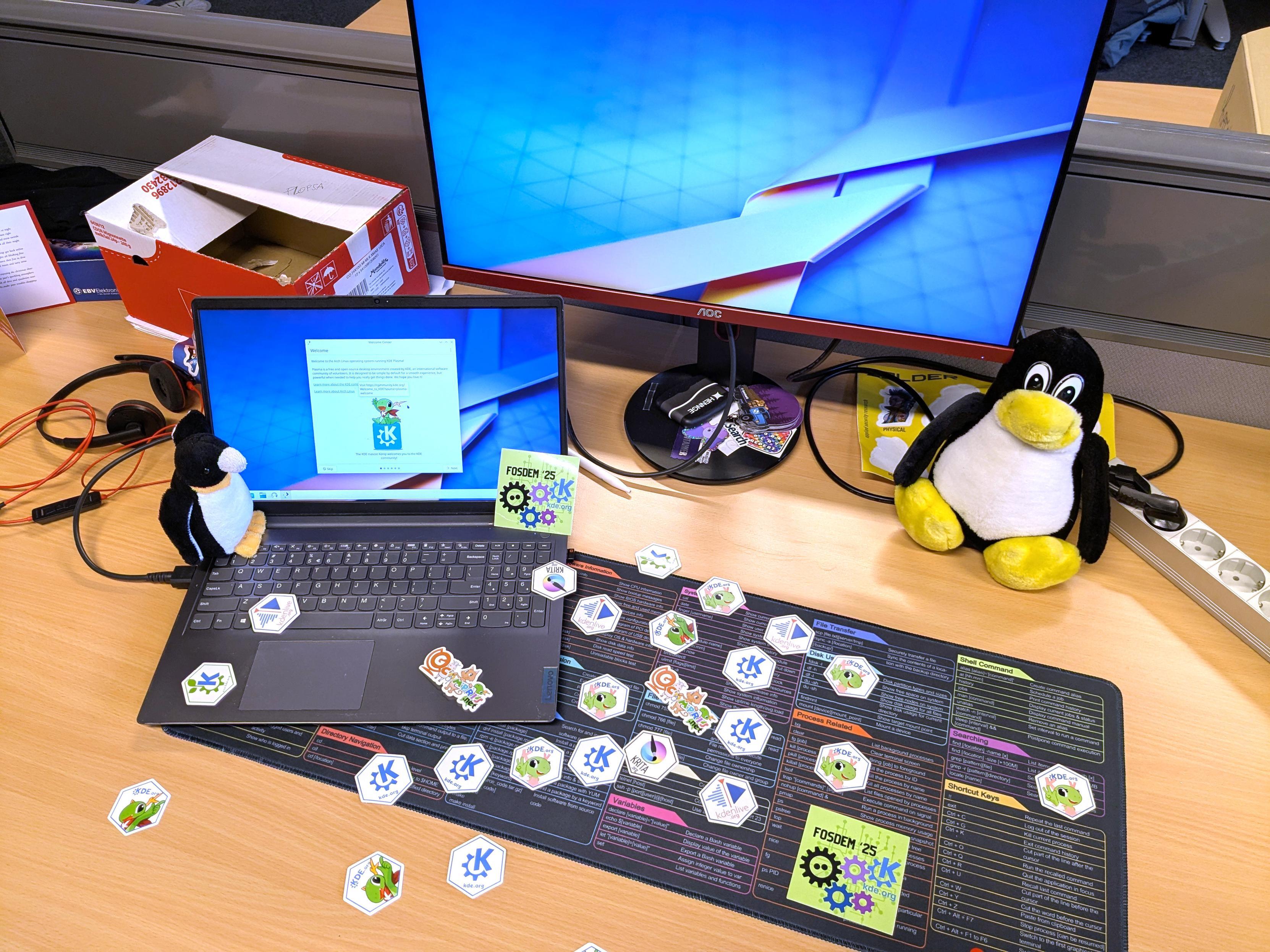 A desk with a laptop and a monitor, both showing KDE's Plasma desktop. Stickers with logos of KDE, Kdenlive, Krita, Gcompris and pictures of Konqi and Katie lie scattered around. On either side are two plushy penguins.